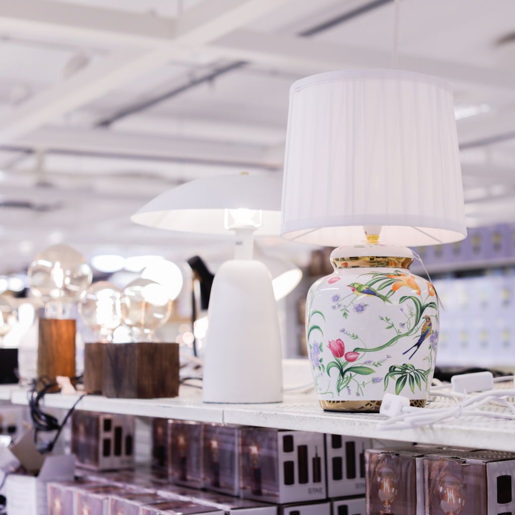 Lampa i porslin med blommönster står på en hylla i en belysningsbutik, omgiven av moderna lampor och glödlampor.