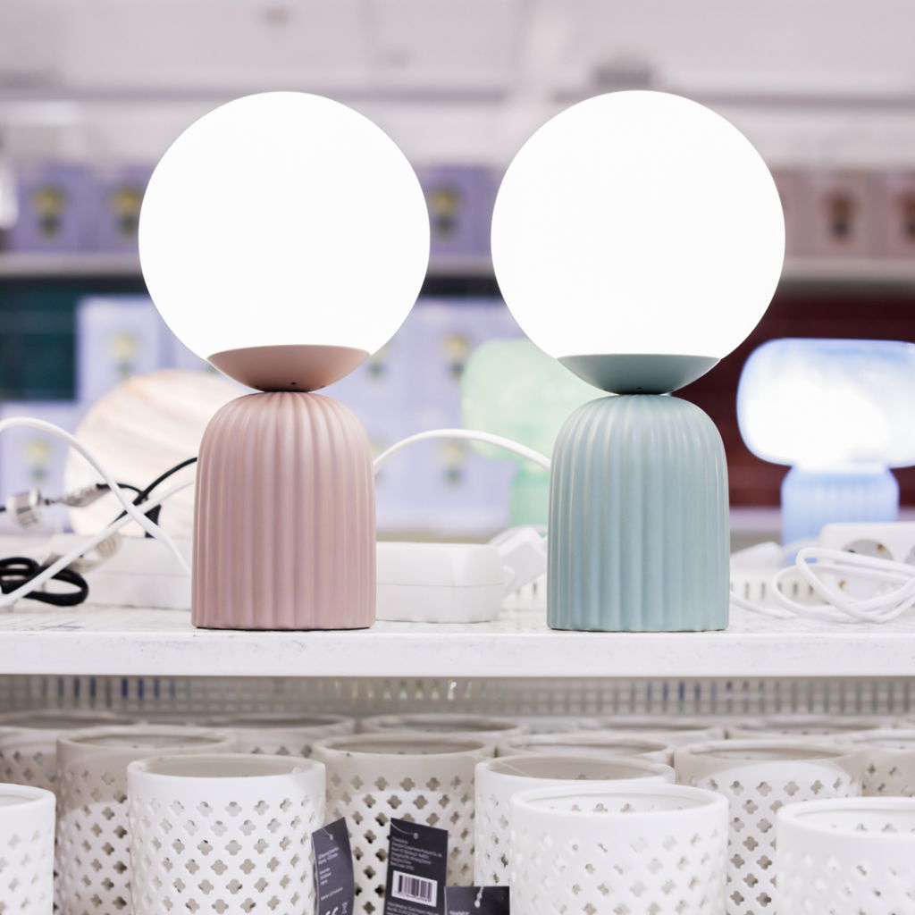 Två moderna lampskärmar i pastellfärger, rosa och blå, med runda glödlampor, placerade på en hylla bland andra dekorativa föremål.