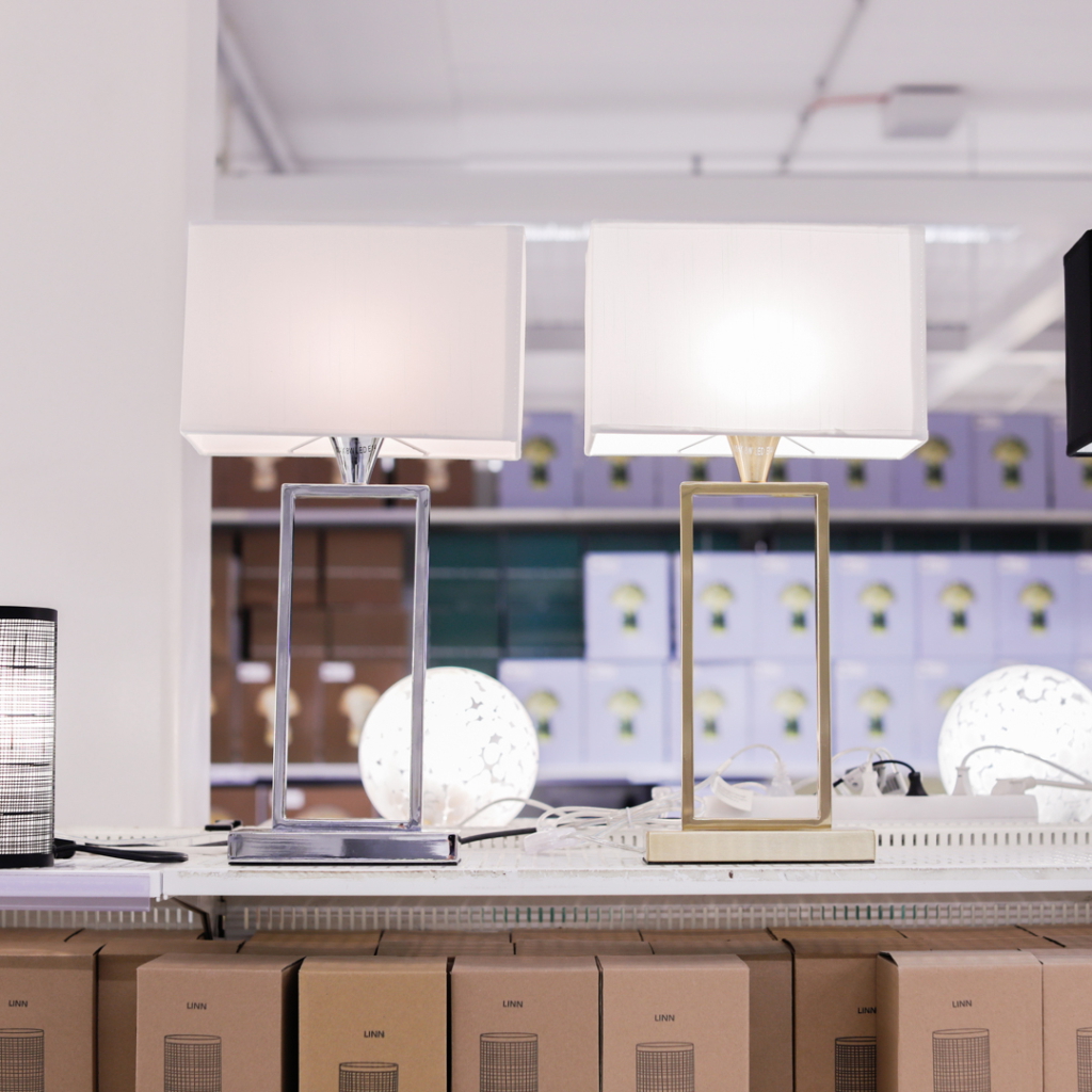 Två moderna bordslampor i silver och guld med vita lampskärmar, placerade på en hylla med förpackningar i bakgrunden.