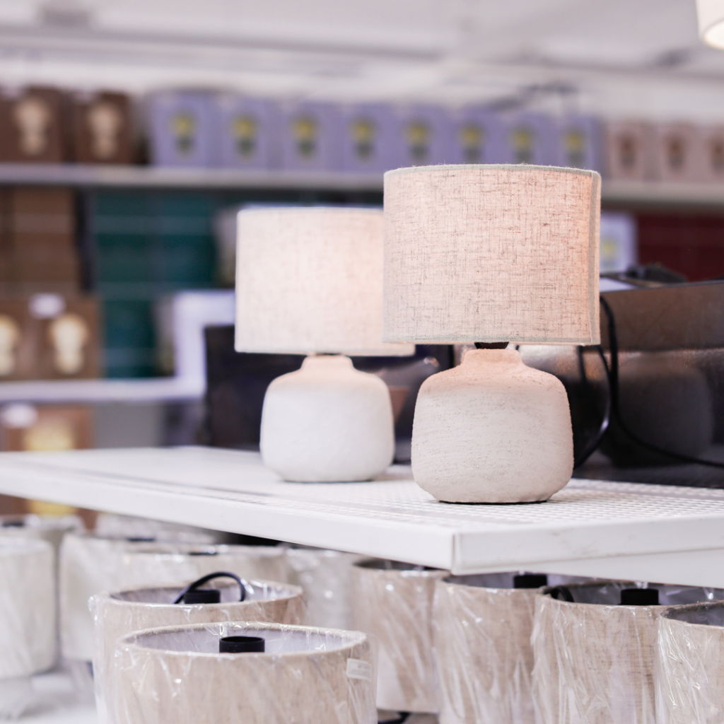 Inredning med moderna bordslampor i en butik, med fokus på lampor med vita skärmar och keramiska baser. Bakgrunden visar förpackade lampor och dekorativa föremål.
