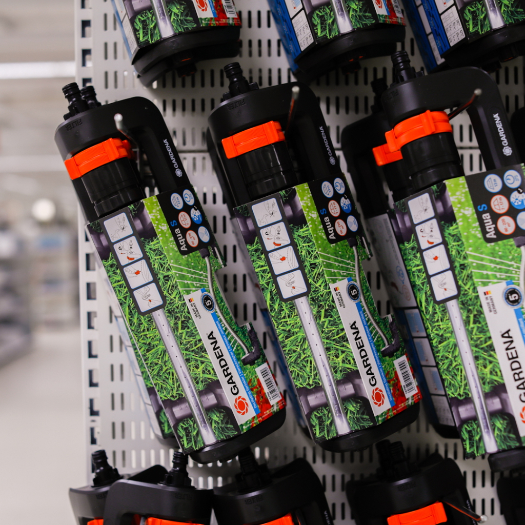 Alt-text: Gardena Aqua S vattenspruta i en butik, med tydliga instruktionssymboler och grönsaksdesign på förpackningen, hängande på en hylla.