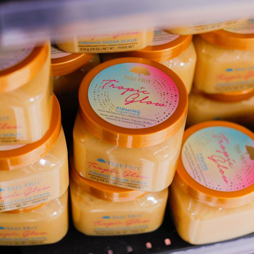 Burkar med Tree Hut Tropic Glow firmande sockerpeeling, staplade i en hylla. Produkten är designad för att ge huden en strålande och fast känsla med naturliga ingredienser.