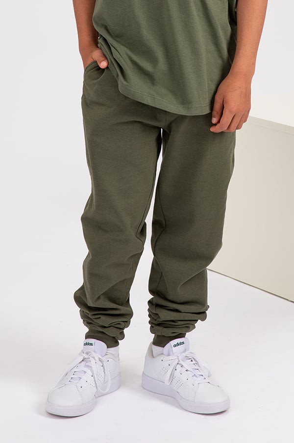 Barn i olivgröna byxor och vita sneakers från Adidas, poserar framför en neutral bakgrund. Byxorna har en avslappnad passform och är perfekta för vardagsbruk.