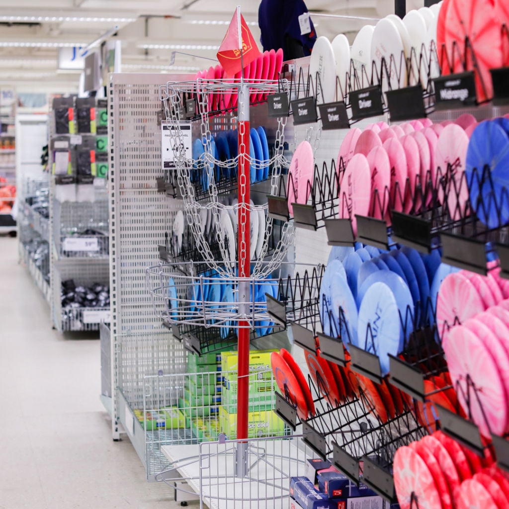 Discgolfutrustning i en sportbutik med en uppsättning färgglada discar i olika nyanser, inklusive rosa, blå och vita, samt en discgolfkorg i mitten av hyllan.