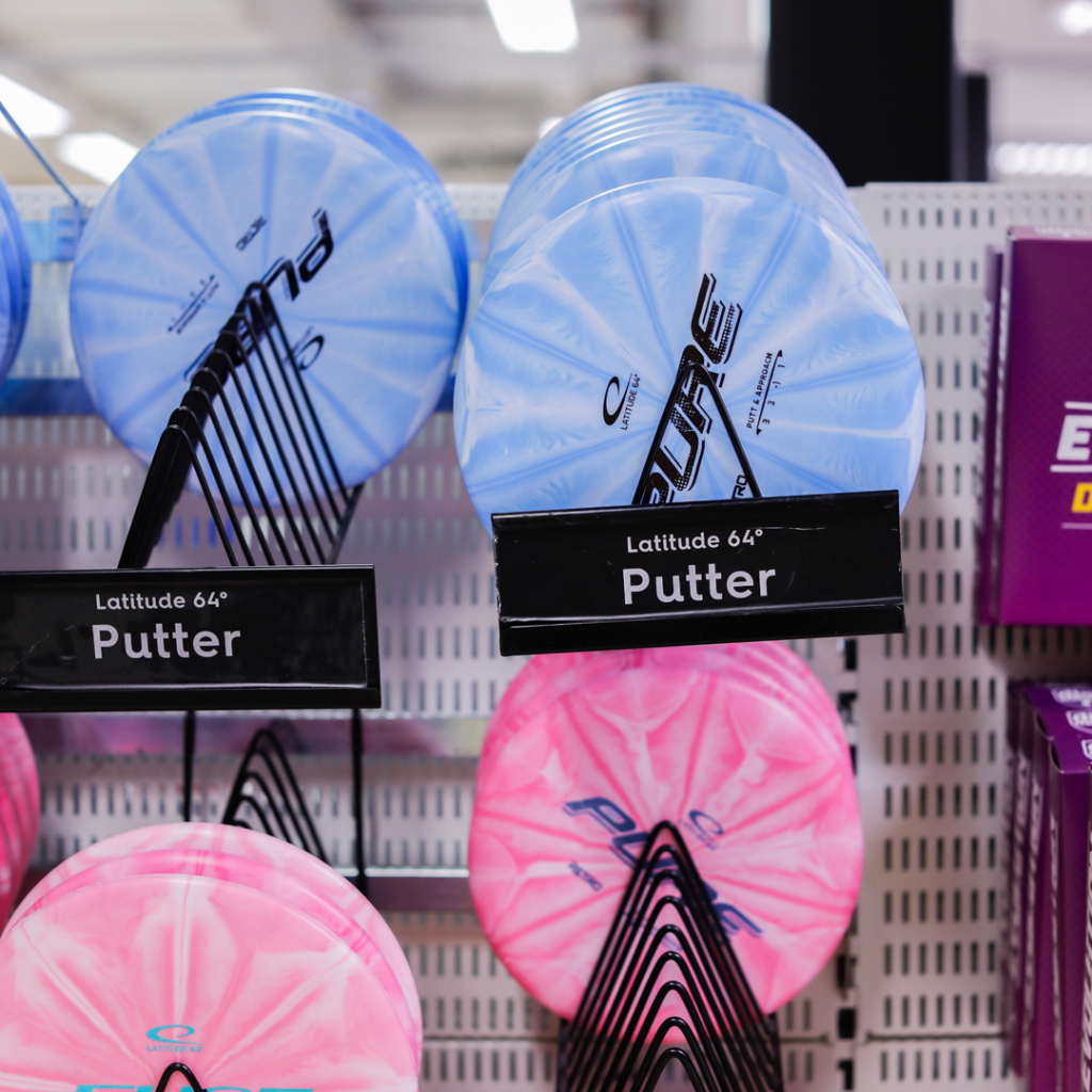 Flera putters från Latitude 64° i blått och rosa, utställda på hylla i sportbutik, med tydliga etiketter som visar produktnamnet.
