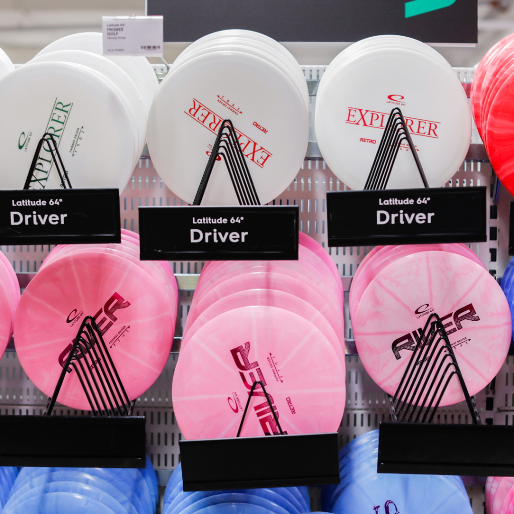 Flera discgolfskivor av märket Latitude 64° på en hylla, inklusive modellerna Explorer och River i olika färger som vitt, rosa och blått.