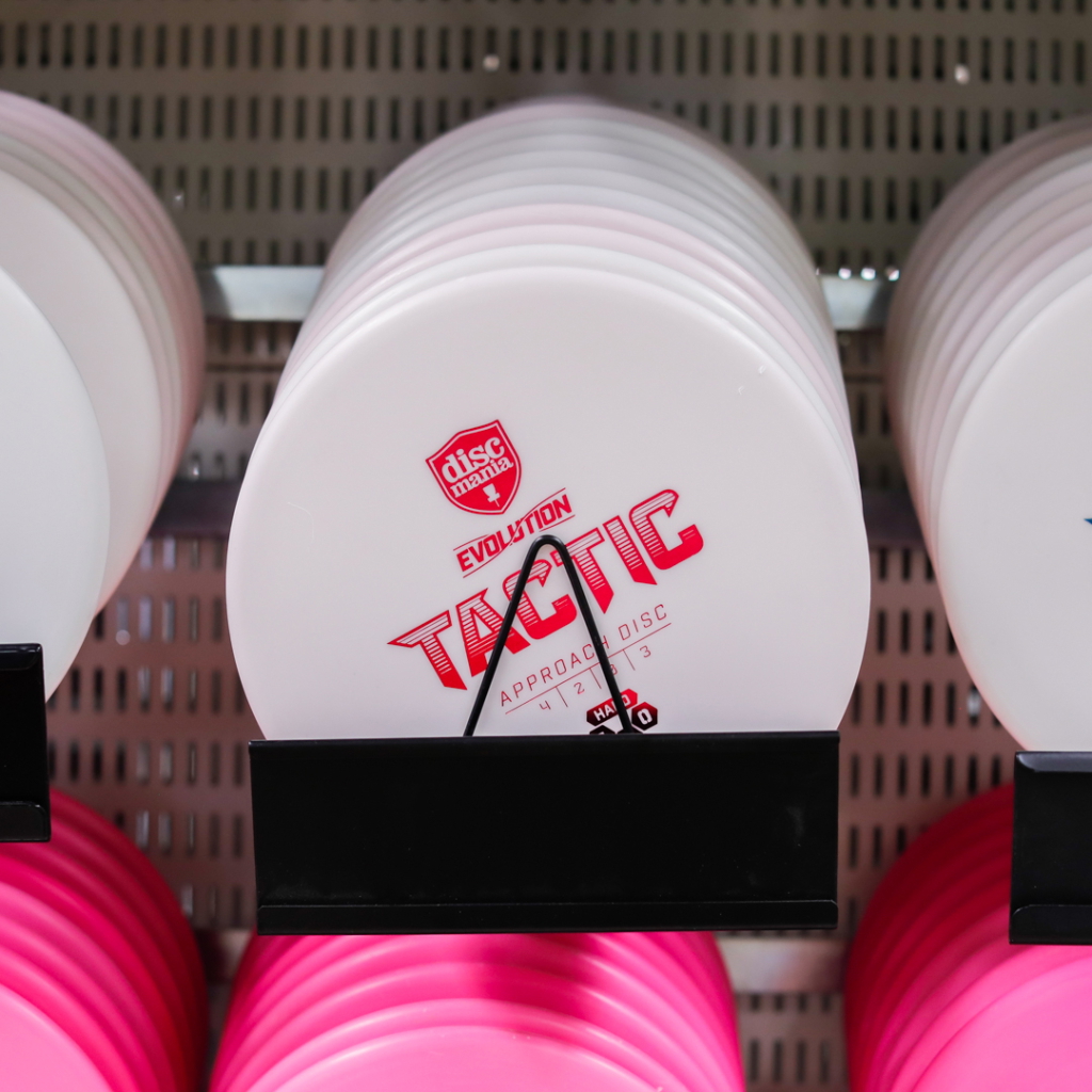 Vit discgolfskiva från Discmania, modell Tactic, med röd text och design. Skivan är avsedd för närspel och visas i en hylla tillsammans med andra färgade discar.
