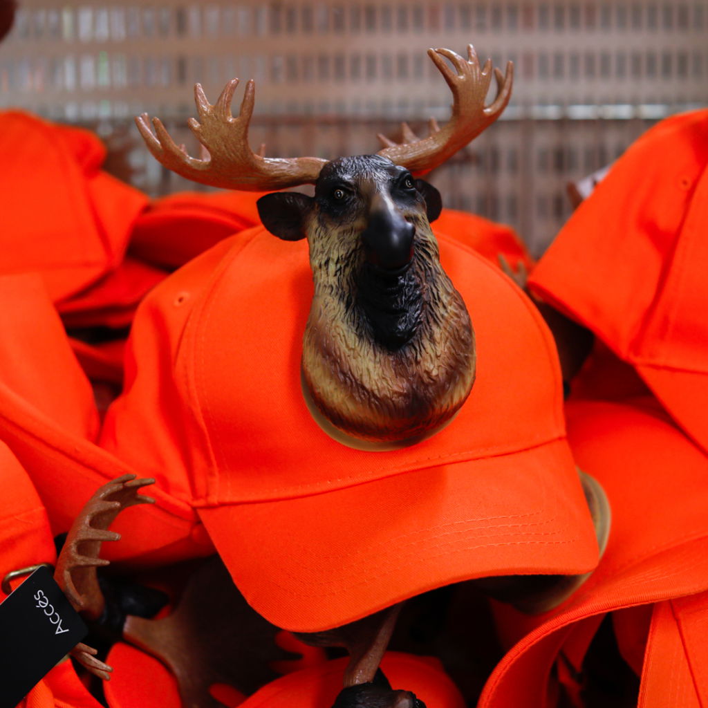 En orange keps med en plastfigur av en älg med horn som dekoration, omgiven av flera andra orangea kepsar. Perfekt för jakt eller utomhusaktiviteter.