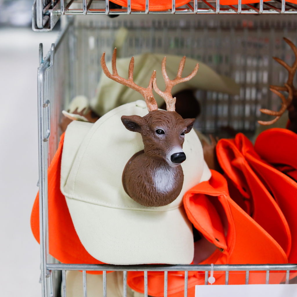 En hylla med kepsar, inklusive en beige keps med en plastdekor av ett rådjurshuvud med horn, omgiven av orange kepsar.