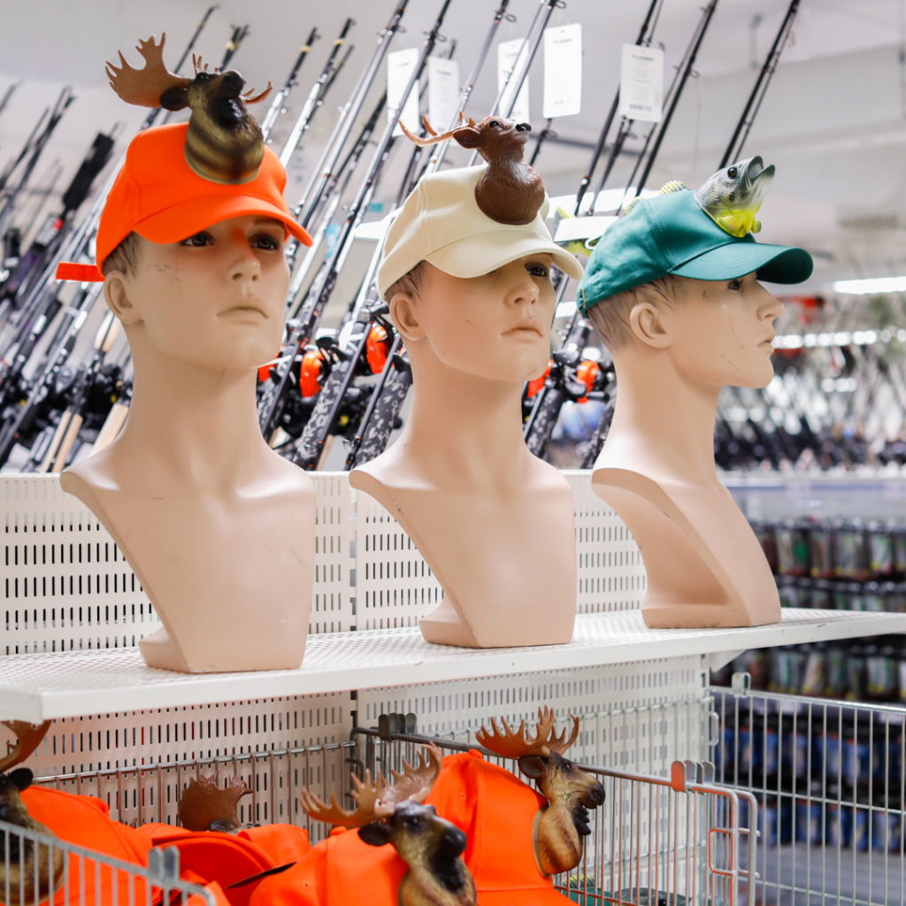 Tre mannekänger med olika kepsar prydda med leksaksdjur, inklusive en älg och en fisk, i en sportbutik med fiskeutrustning i bakgrunden.