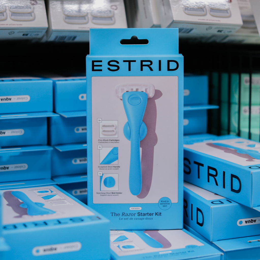 Förpackning av Estrid rakningskit med en blå rakapparat och tillbehör, inklusive fembladiga rakblad och en matchande väggfäste, i en butiksmiljö.