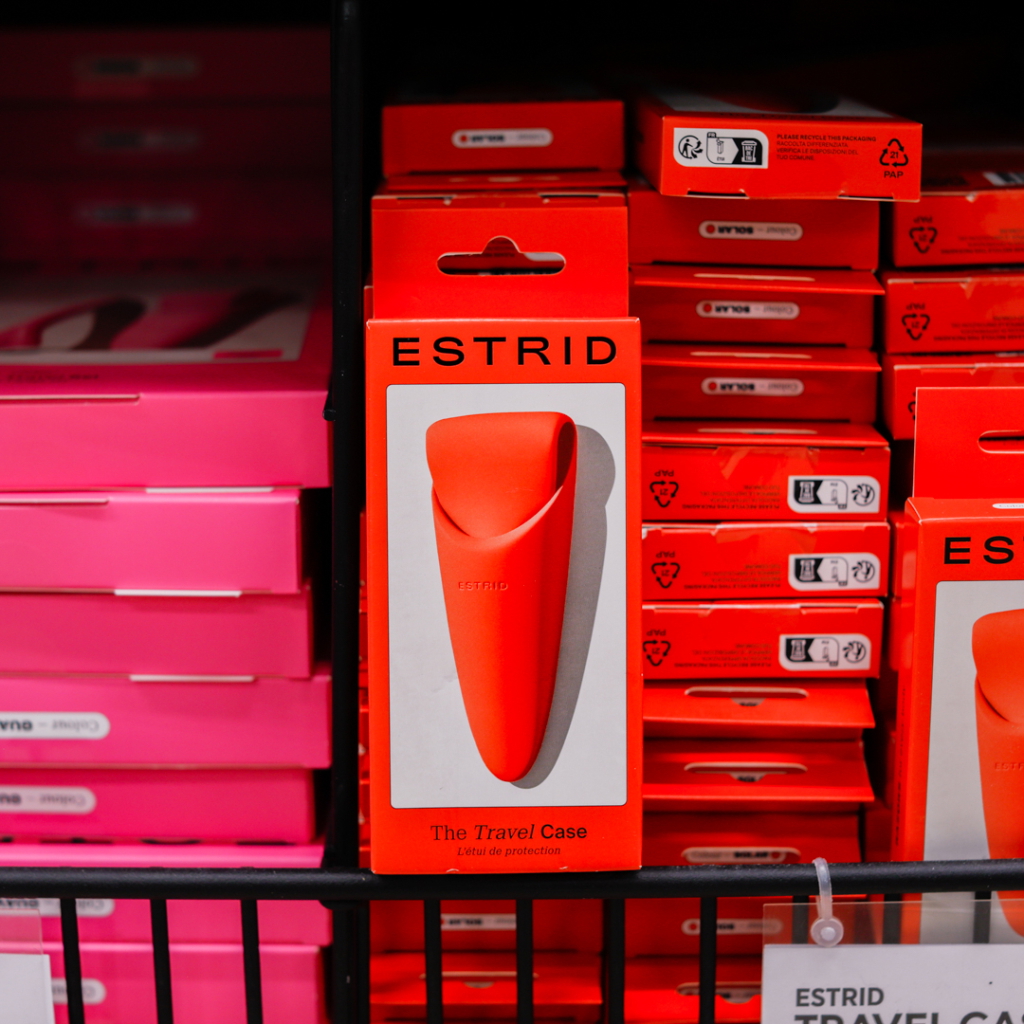 Röd reseförpackning från ESTRID, designad för att skydda och förvara personliga hygienprodukter, placerad bland andra förpackningar i en butik.