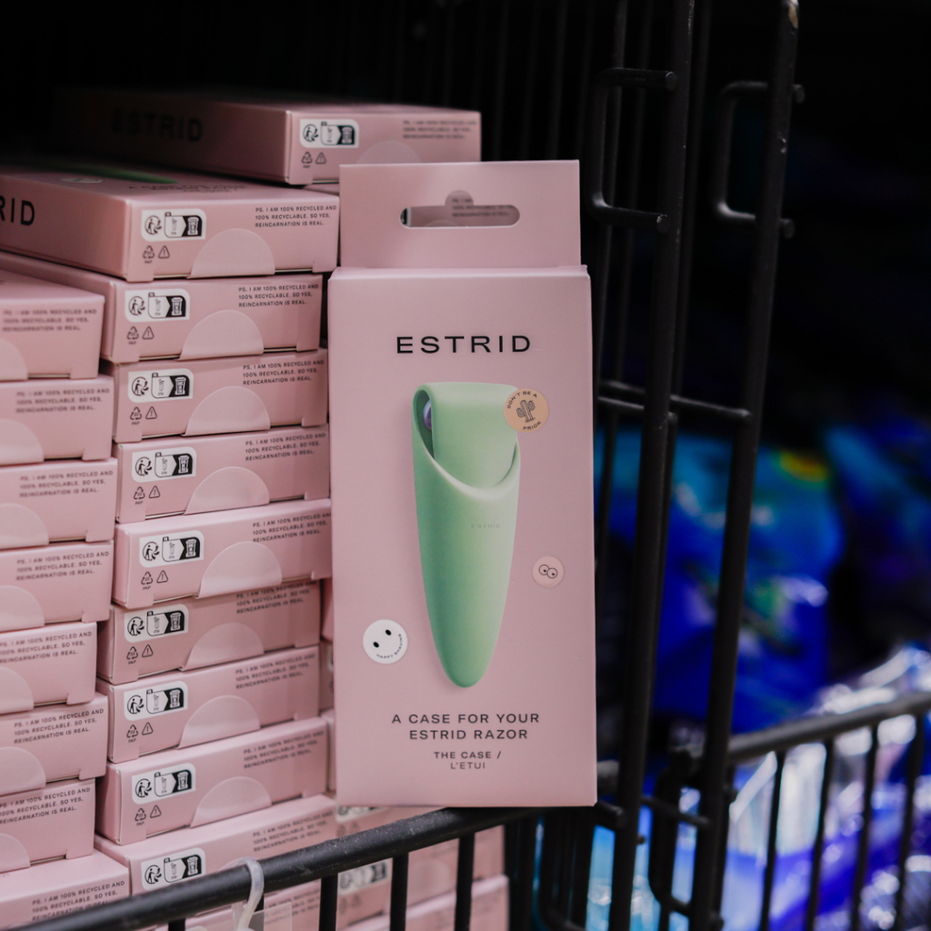 Grön förpackning för ESTRID-rakhyvelnsetui framför en hylla med rosa lådor, designad för att skydda och förvara rakhyveln på ett stiligt sätt.