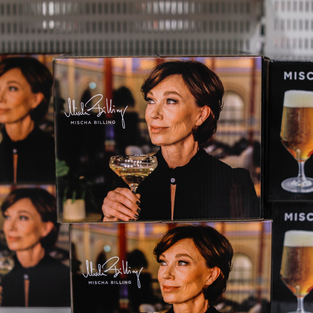 Kvinna med kort brunt hår håller en cocktail och poserar framför en bakgrund med dämpad belysning, på förpackningen för Mischa Billing-produkter.