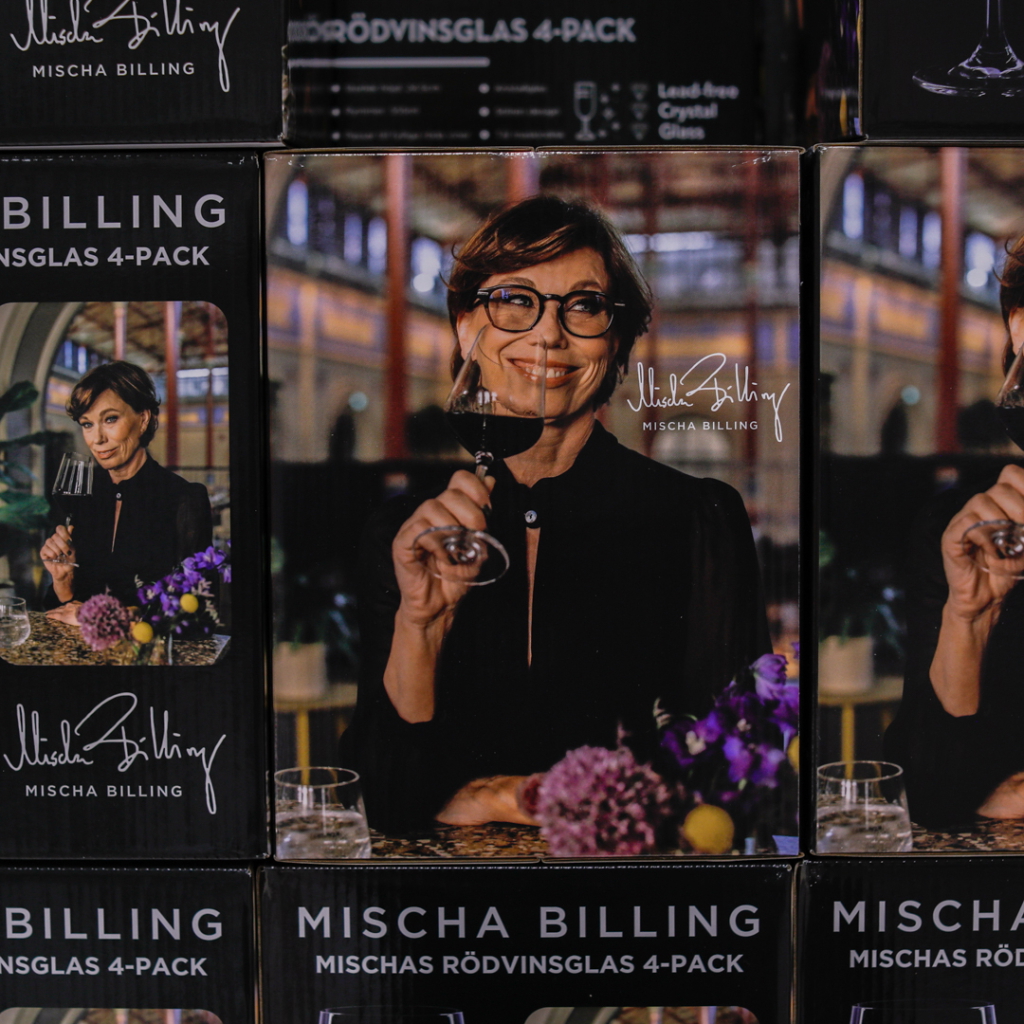 Förpackning av Mischa Billings röda vinglas i 4-pack, med en kvinna som håller ett vinglas och ler. Bakgrunden har blommor och ett stiligt inredningsarrangemang.