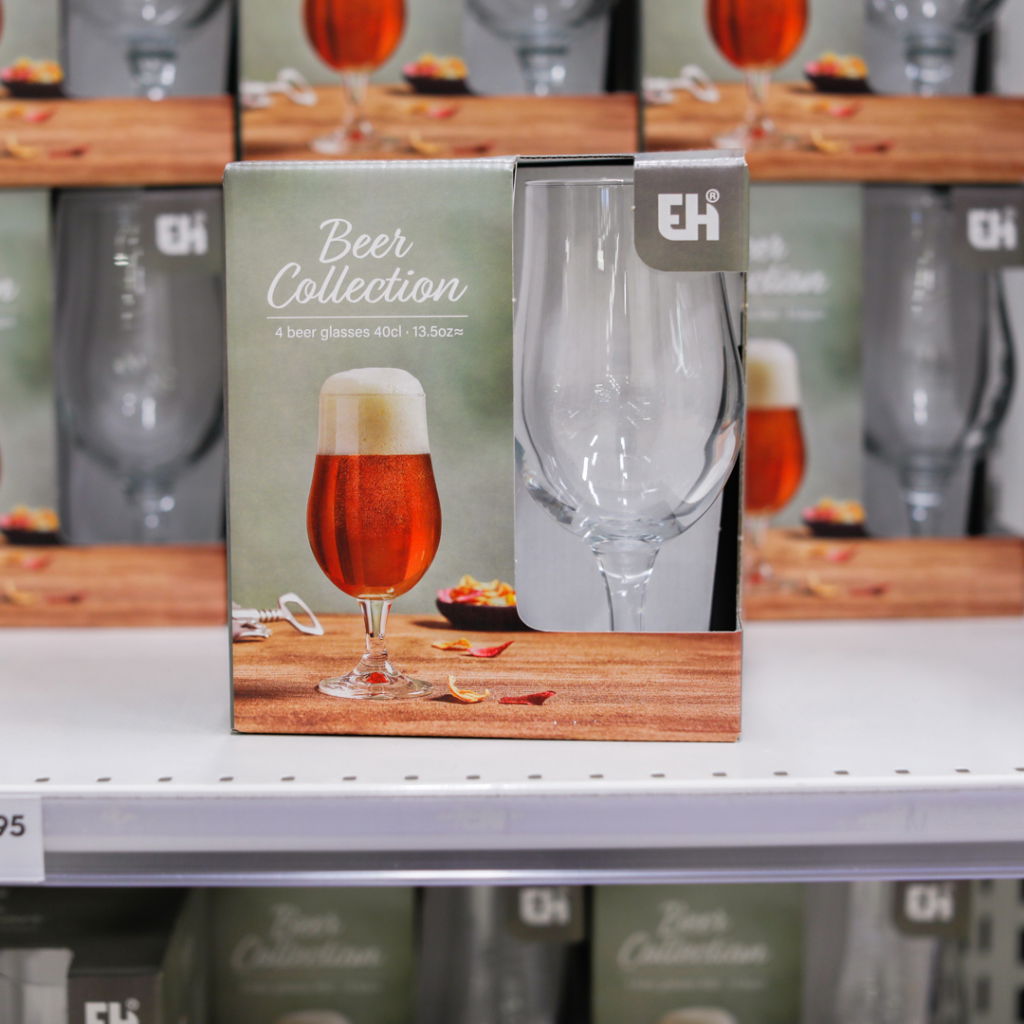 Förpackning med fyra ölglas i glas, 40 cl, med en bild av ett glas öl och en öppen korkskruv. Bakgrunden visar flera liknande förpackningar i en butik.