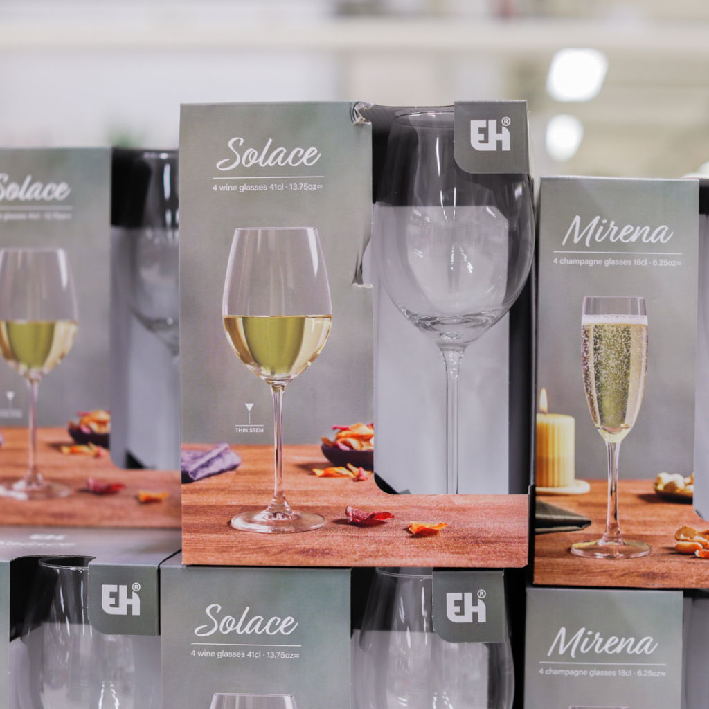 Förpackningar med vinglas och champagneglas från EH, inklusive serien Solace med fyra vinglas (41 cl) och Mirena med fyra champagneglas (18 cl), placerade på en träyta med dekorativa detaljer.