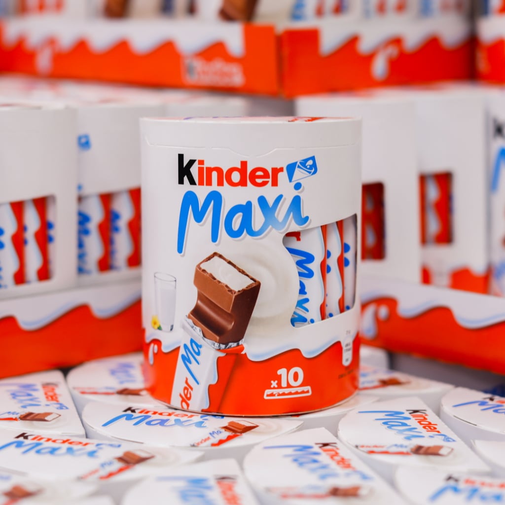 Förpackning av Kinder Maxi med tio chokladstänger, omgiven av flera liknande förpackningar. Chokladen har en mjuk fyllning och är dekorerad med röd och vit design.