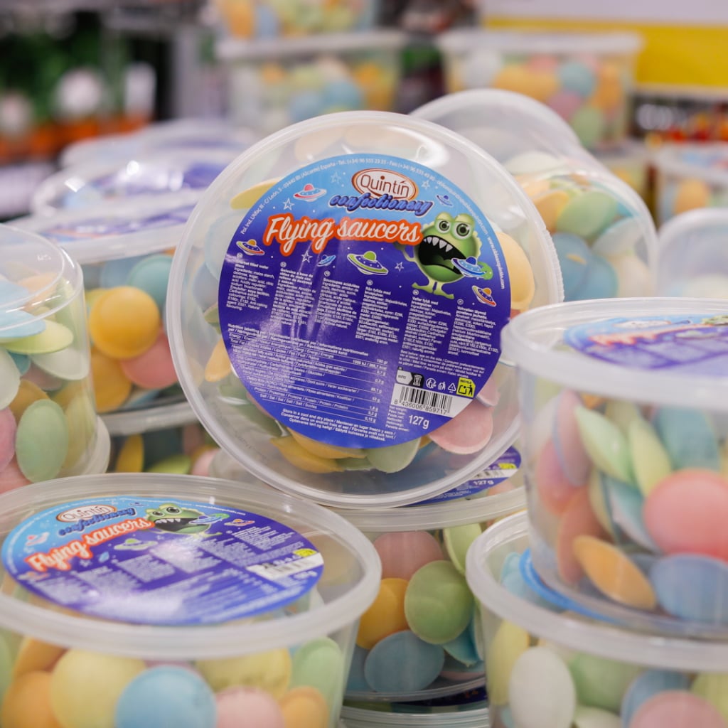 Färgglada Flying Saucer-godisar i plastbehållare, märkta med "Quintin" och en tecknad utomjording. Godisarna är i olika pastellfärger och ligger staplade i en butiksmiljö. Varje behållare innehåller 127 gram av dessa sötsaker.