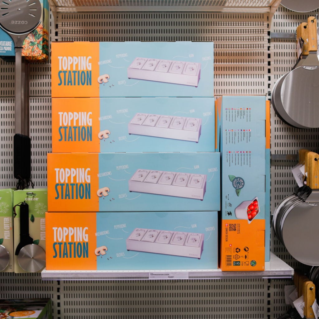 Förpackningar av Topping Station på en hylla, med olika pålägg som pepperoni, svamp, oliver, skinka och lök tydligt angivna. Produkten är avsedd för att enkelt servera pizza-tillbehör.