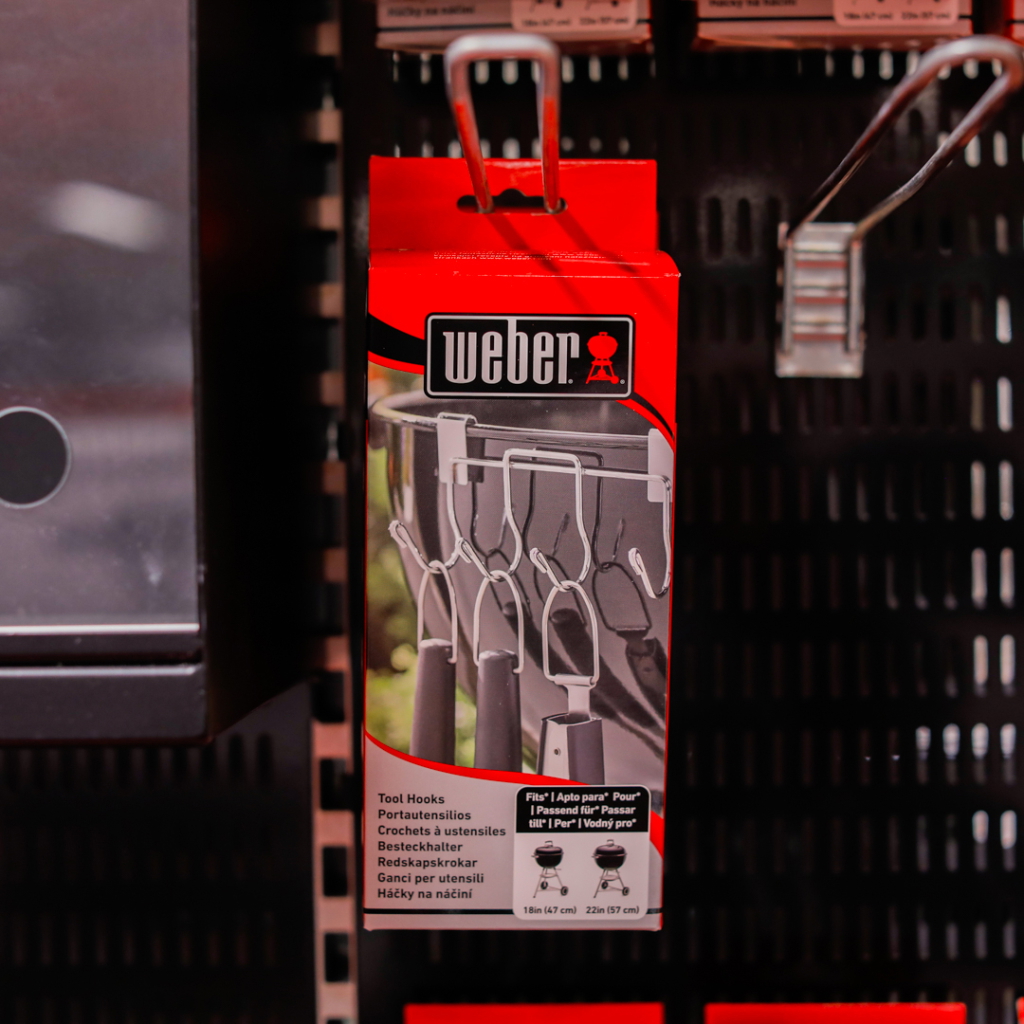 Förpackning med Weber verktygskrokar, designad för att hänga grillredskap, placerad på en svart vägg.