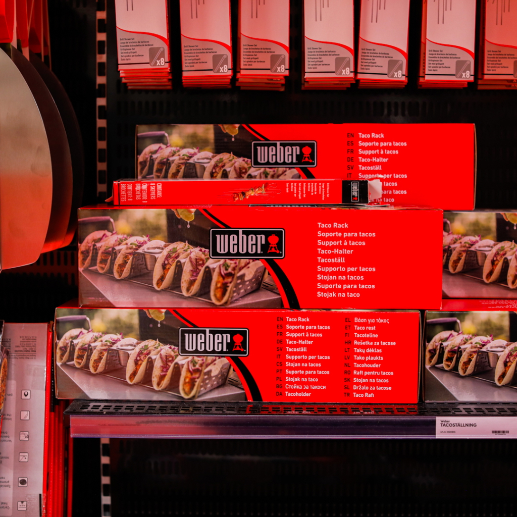 Förpackningar med Weber Taco Rack, designad för att hålla tacos på plats under matlagning. Produkten visas på en hylla i en butik, med flera språk på förpackningen.