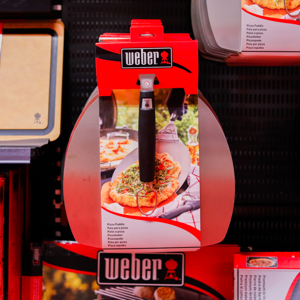 Bild av en Weber pizzaspade som hänger i butik, med svart handtag och metallblad, perfekt för att lyfta och servera pizza.
