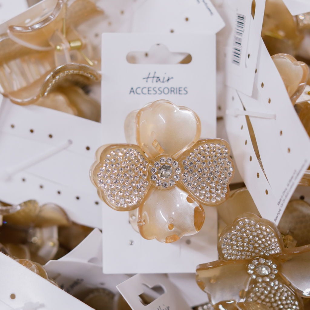 Håraccessoarer med en blomformad hårklämma i beige och strassdetaljer, omgivna av flera andra liknande produkter på en vit bakgrund.