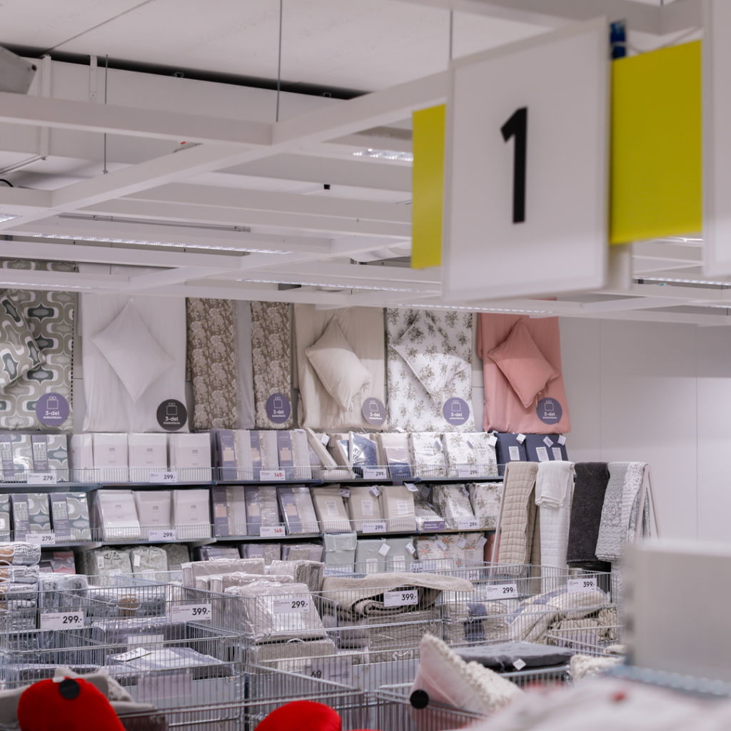 Bild av en heminredningsavdelning i en butik med sängkläder, kuddar och plädar på hyllor. Priserna är tydligt märkta på produkterna.