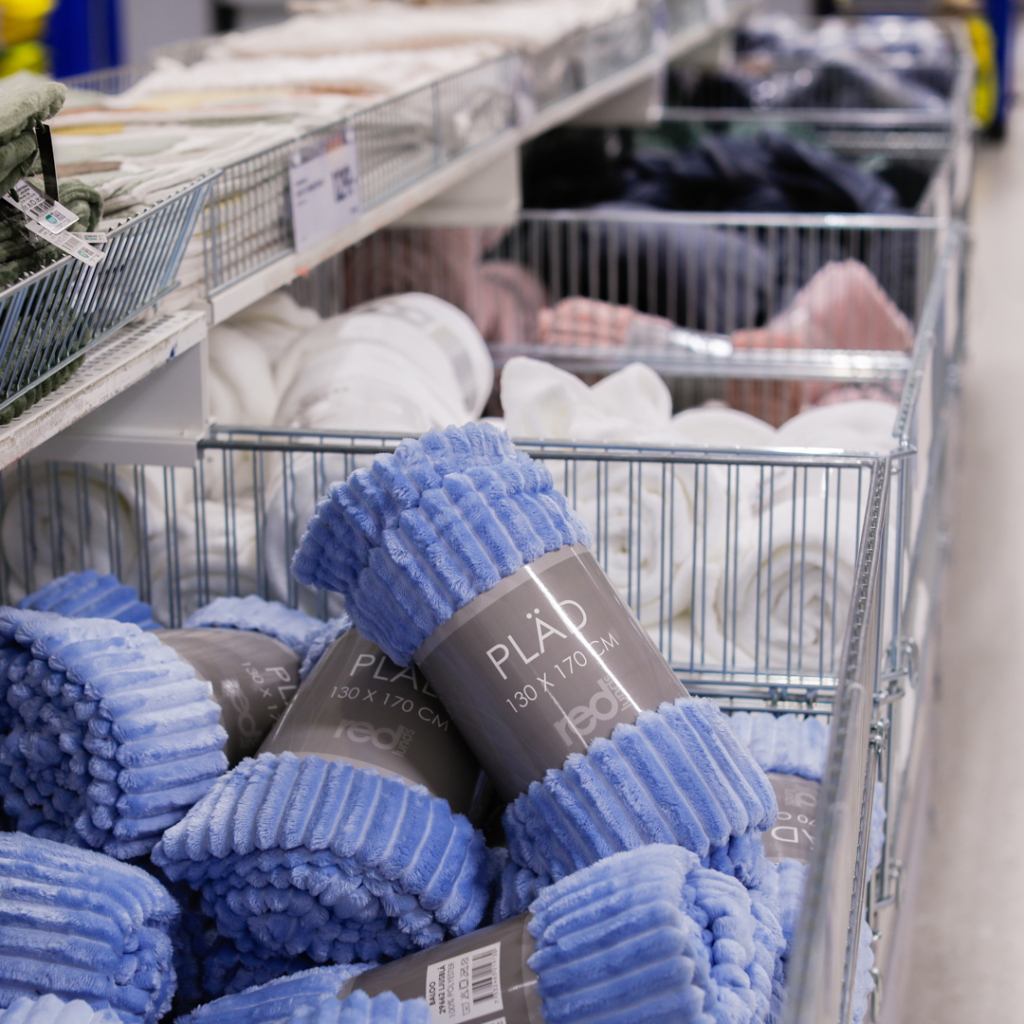 Blå plädar i en butik, förpackade i rullar med måtten 130 x 170 cm, placerade i korgar tillsammans med andra textilier som handdukar och filtar.