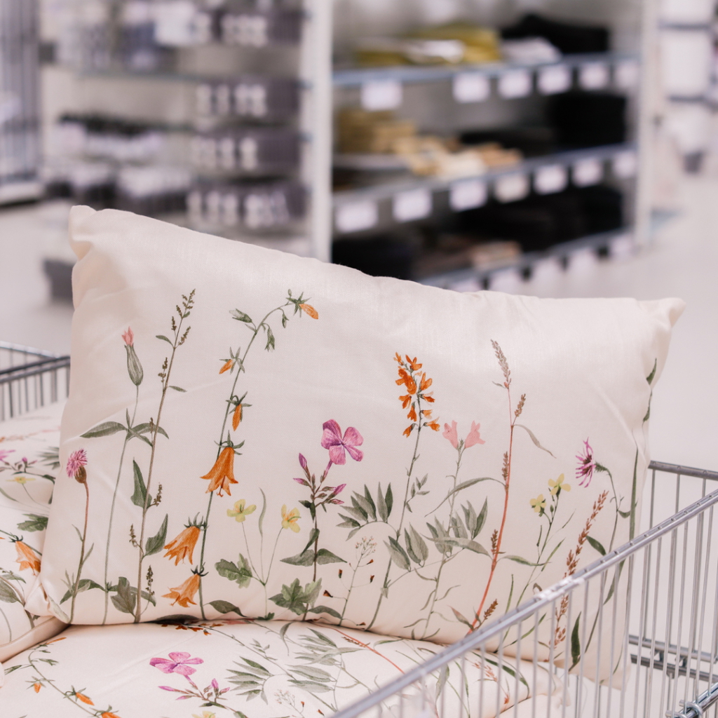 Dekorativ kudde med blommönster i pastellfärger, placerad i en kundvagn i en heminredningsbutik. Bakgrunden visar hyllor med olika textilier och inredningsprodukter.
