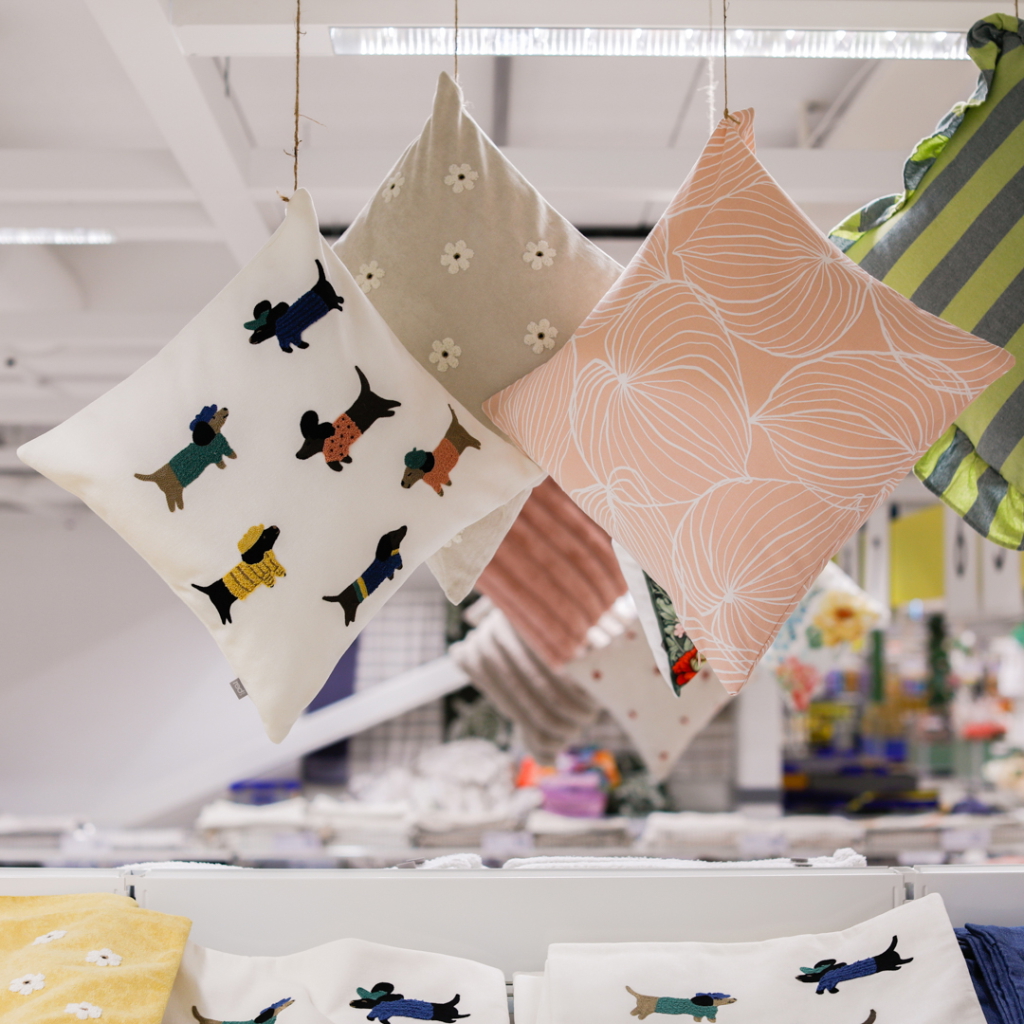 Färgglada kuddar med hundmotiv hänger i en butik, omgivna av olika textilier och dekorationer. Kuddarna har olika mönster, inklusive blommor och abstrakta former, vilket ger en livlig och inbjudande atmosfär.