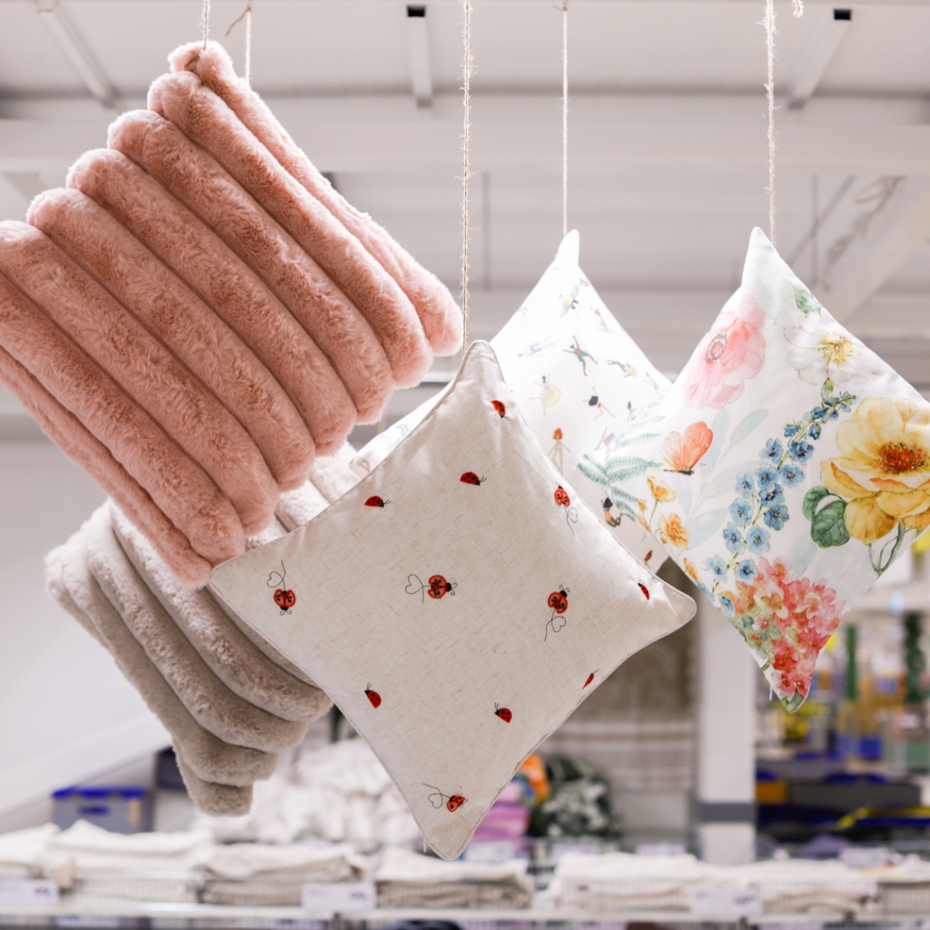 Flera dekorativa kuddar hängande i en butik, inklusive en rosa plyschkudde och mönstrade kuddar med blommor och insekter, som ger en hemtrevlig känsla.
