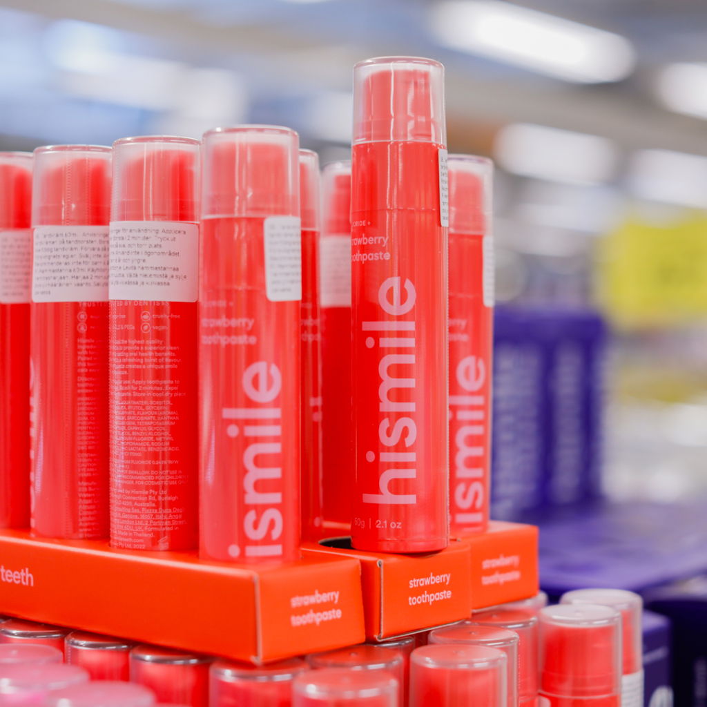 Röd tandkräm från Hismile med smak av jordgubbe, placerad på hyllor i en butik, omgiven av flera andra tandkrämförpackningar.