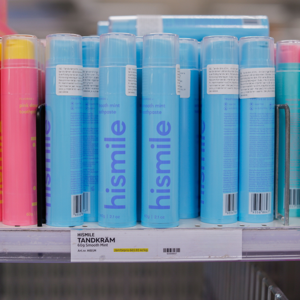 Bild av flera flaskor med tandkräm från märket Hismile, inklusive smooth mint och pink donut varianter, placerade på en hylla i en butik. Produkten är förpackad i färgglada rör och har etiketter med information om innehåll och pris.