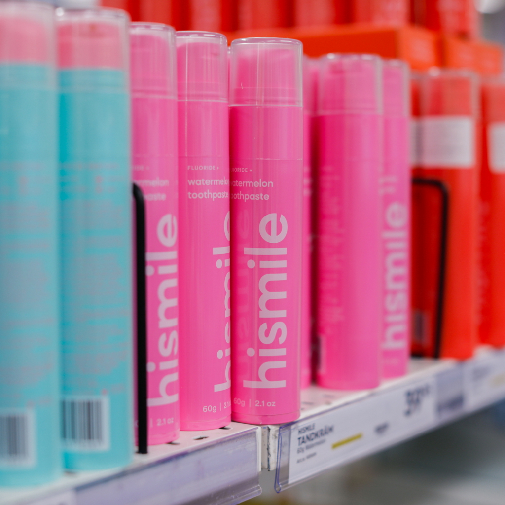 Flaskor med tandkräm från märket Hismile i olika färger, inklusive rosa och blå, på en hylla i en butik. Produkten är vattenmelon-smakande och innehåller fluor.