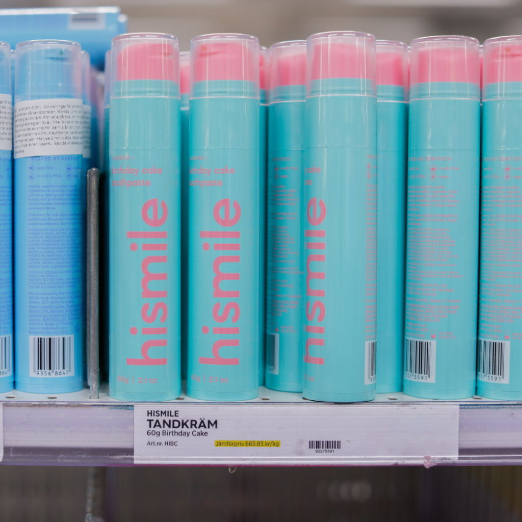 Hismile tandkräm med smak av födelsedagstårta, 60g, placerad på hyllan i en butik. Förpackningen är ljusblå med rosa text, vilket gör den iögonfallande.