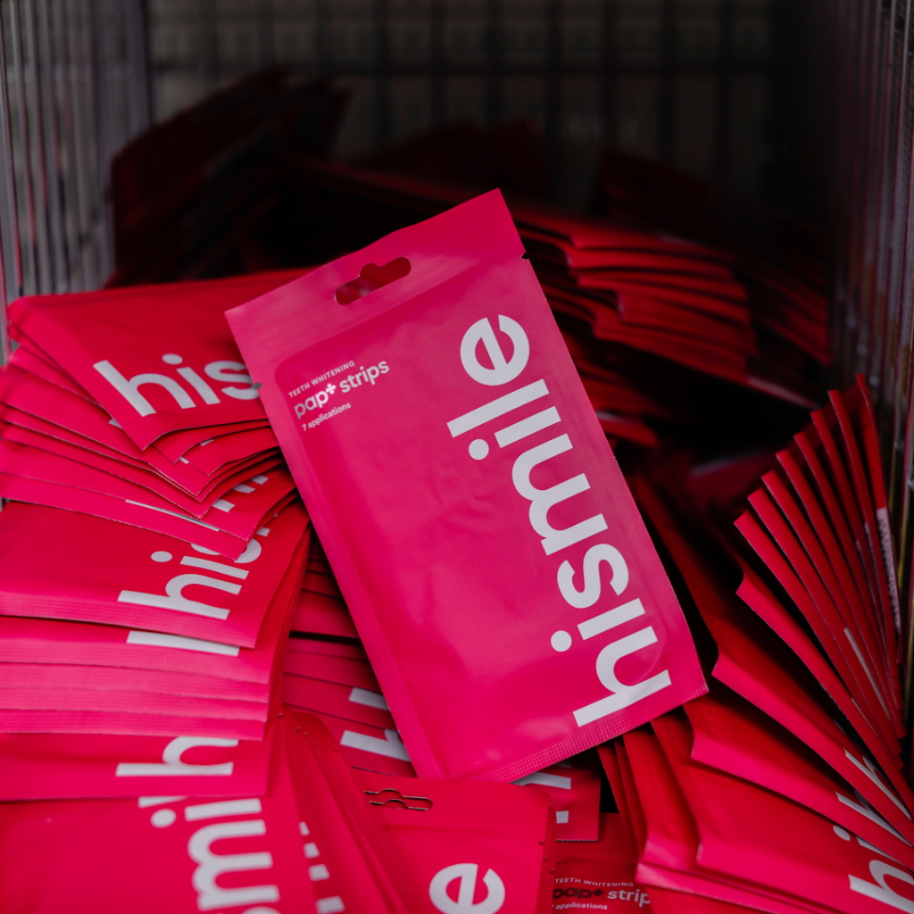 Förpackningar med tandblekningsremsor från Hismile i stark rosa färg, staplade i en korg. Varje paket innehåller 7 applikationer.