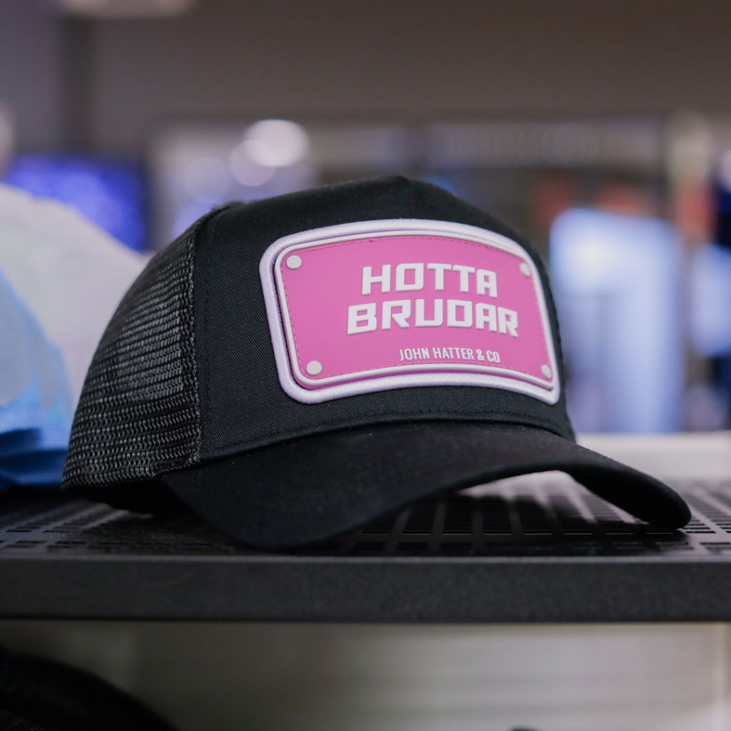Svart keps med rosa emblem som säger "HOTTA BRUDAR" från John Hatter & Co, placerad på en hylla i en butik.