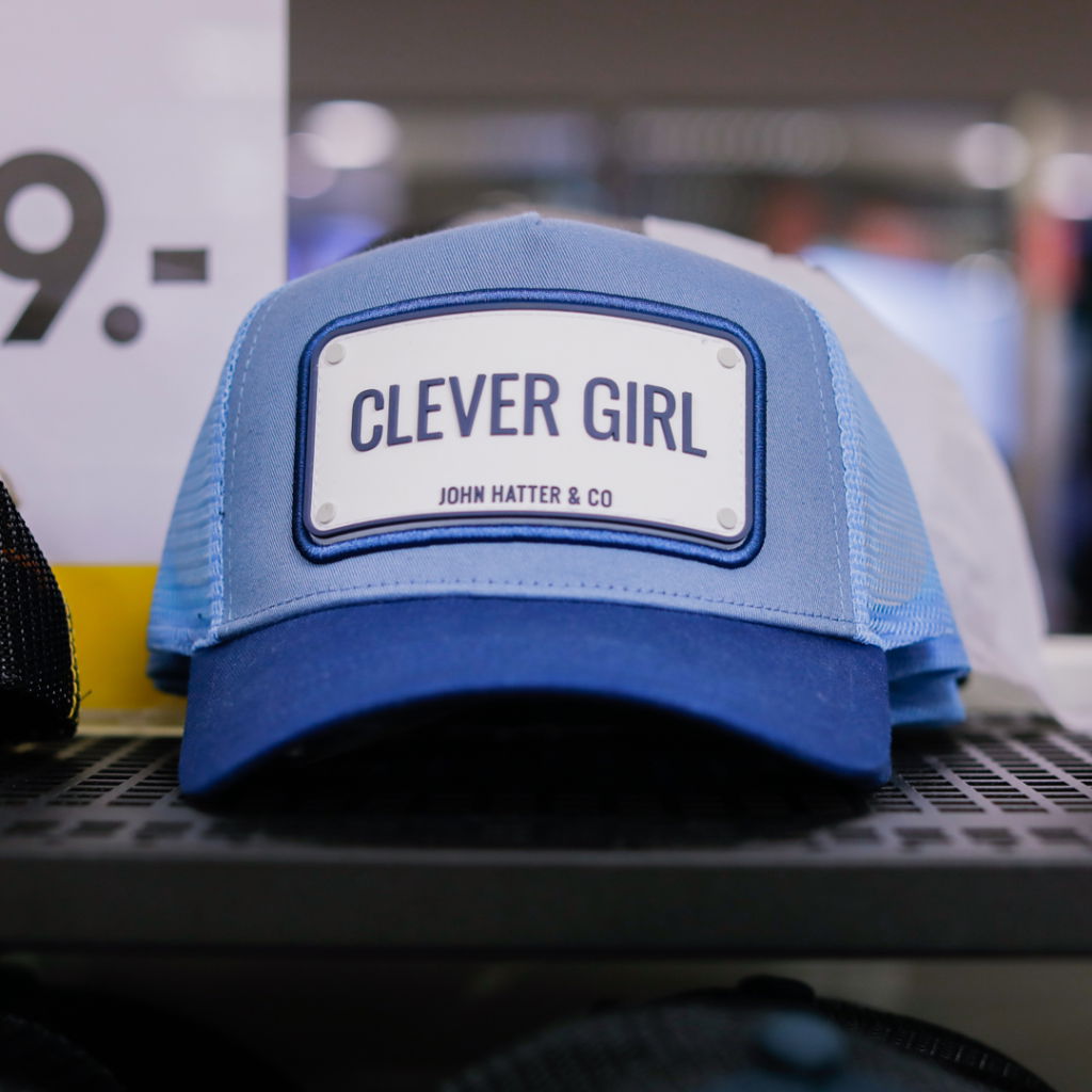 Blå keps med vit skärm och texten "CLEVER GIRL" från John Hatter & Co, placerad på en hylla i en butik.