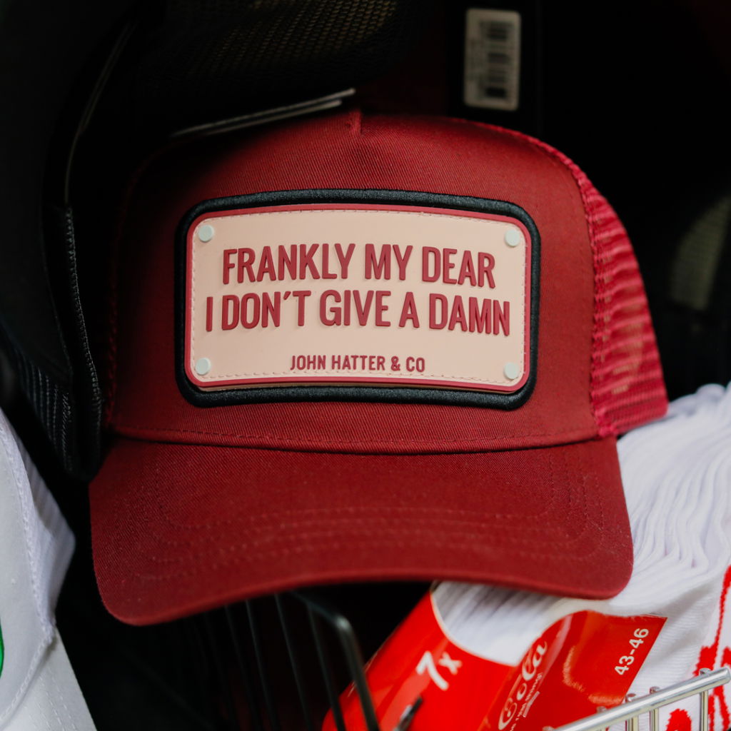 Röd keps med texten "Frankly my dear I don't give a damn" från John Hatter & Co, placerad i en korg tillsammans med vita strumpor och andra klädesplagg.