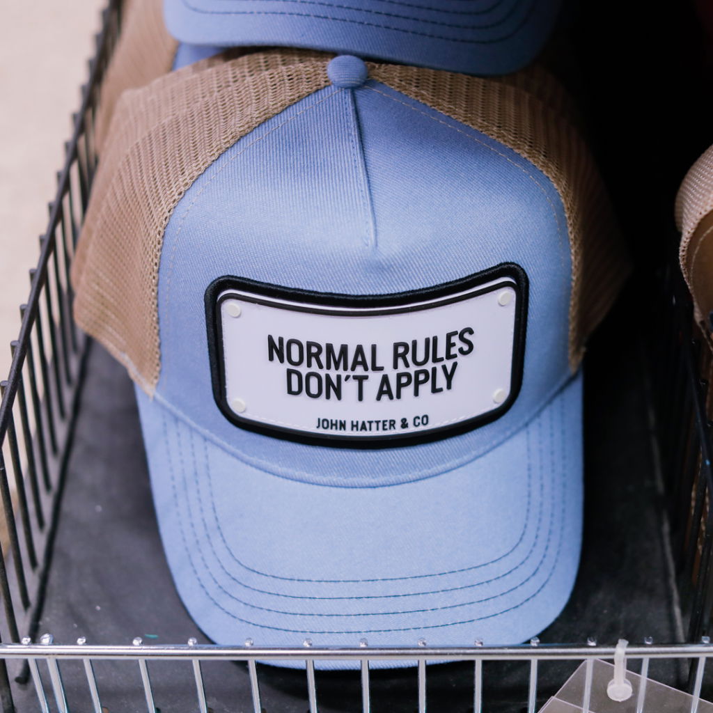 Blå keps med nätbakgrund och texten "Normal Rules Don't Apply" från John Hatter & Co, placerad i en korg.