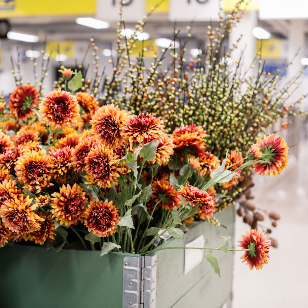 Färgglada konstgjorda blommor i en grön låda, med orange och röda solrosliknande blommor samt grenar med bär, i en modern butiksmiljö.