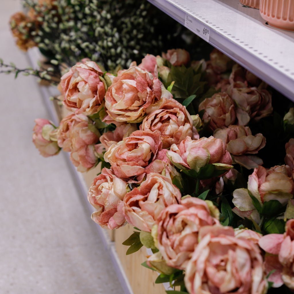 Konstgjorda rosor i ljusrosa nyanser placerade på en hylla i en butik. Blommorna har detaljerade kronblad och gröna bladverk, vilket ger en naturlig känsla. Idealisk för inredning och dekoration.