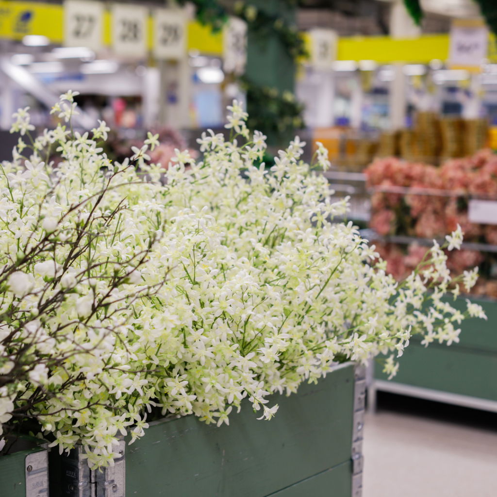 Vita blommor i en grön låda på en blomstermarknad, med oskarpa bakgrunder som visar andra blommor och marknadsaktiviteter.
