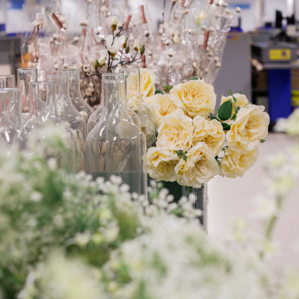 Vackra gula rosor i en floristbutik, omgivna av glasflaskor och dekorativa blommor. Bakgrunden visar fler blommor i olika färger och former, vilket skapar en inspirerande och färgglad atmosfär.