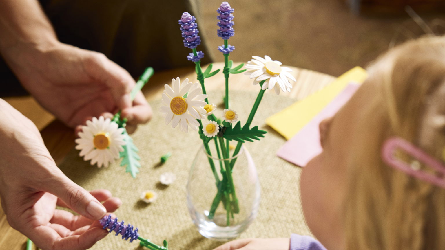 Barn och vuxen skapar blommor av plast, med vita prästkragar och lila lavendel, i en vas på ett bord. Färgglada papper syns i bakgrunden, vilket ger en kreativ och lekfull atmosfär.