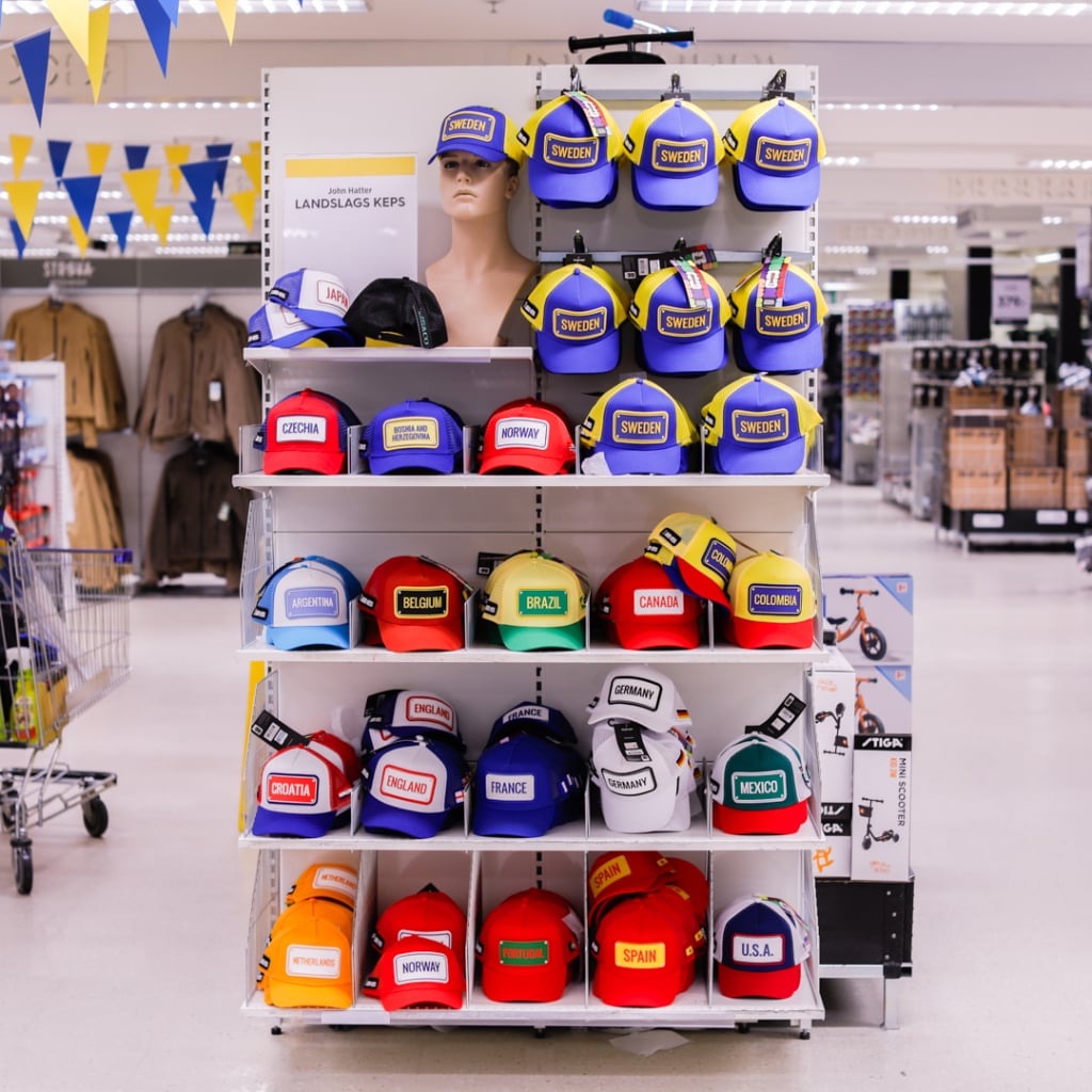 En hylla med färgglada kepsar med olika länders flaggor och namn, inklusive Sverige, Brasilien, Kanada och många fler, i en butiksmiljö.