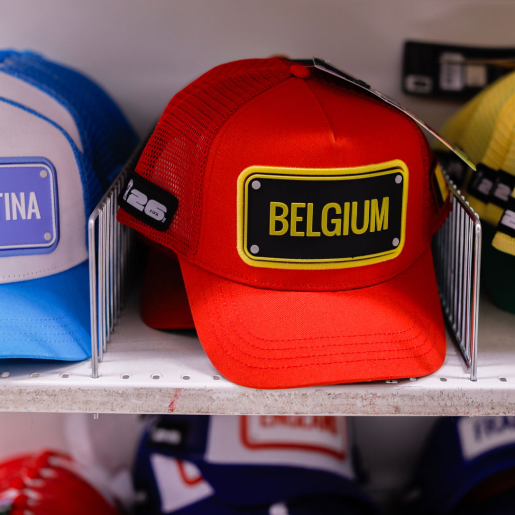 Röd keps med texten "BELGIUM" i gult och svart, placerad bland andra färgglada kepsar i en butik.