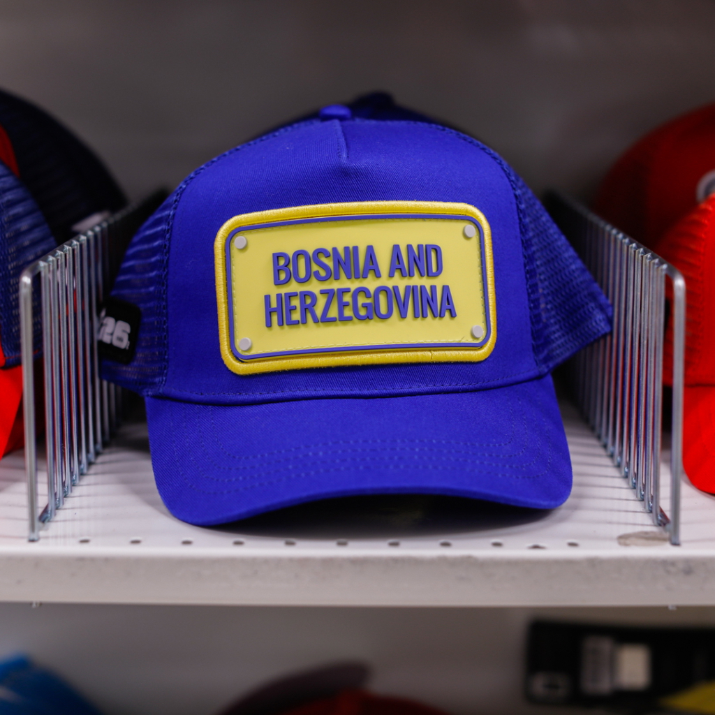Blå keps med texten "Bosnien och Hercegovina" på en gul skylt, placerad på en hylla bland andra kepsar.